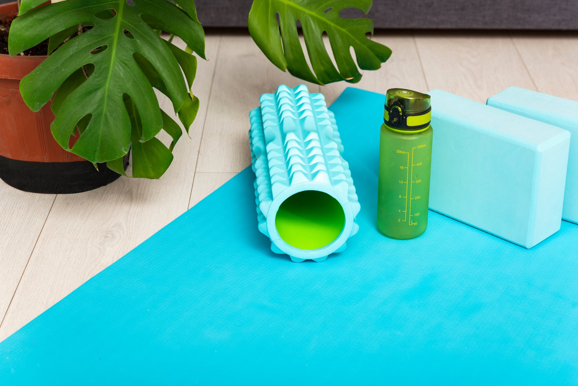 Yoga mat with foam roller, water bottle, and blocks on a light wood floor.